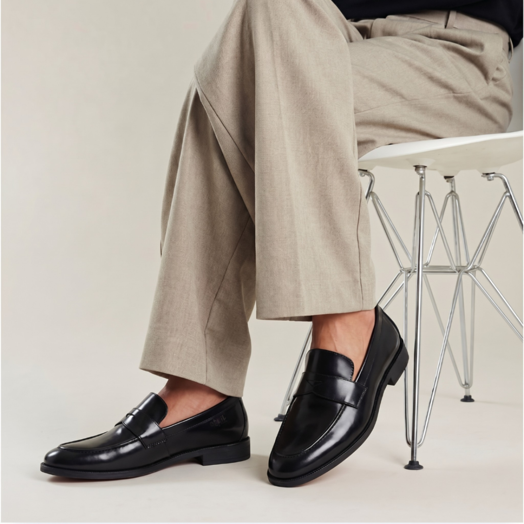 A person seated on a white chair wearing black leather penny loafers with wide-leg beige trousers, photographed in a clean studio setting.