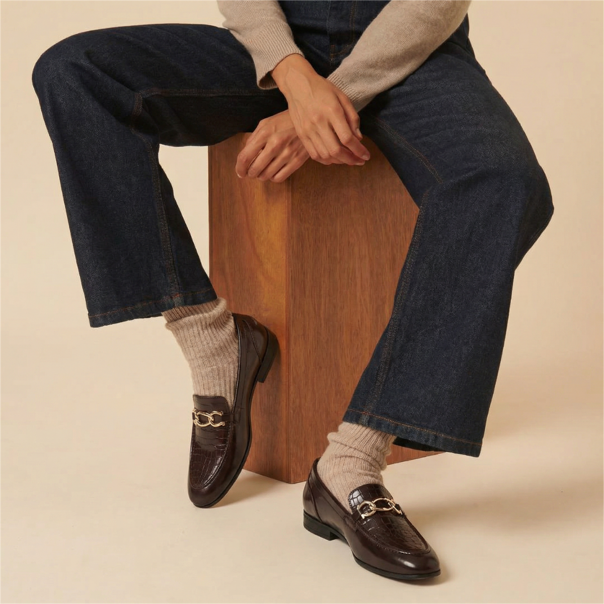 Person wearing dark blue jeans, brown loafers, and beige socks sitting on a wooden block.