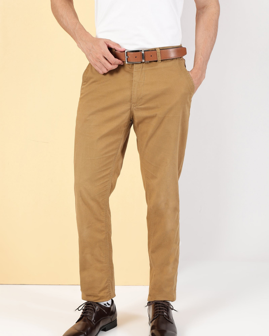 A person wearing slim-fit tan chinos with a brown leather belt, paired with a white t-shirt and dark brown dress shoes, standing against a split beige and white studio background.