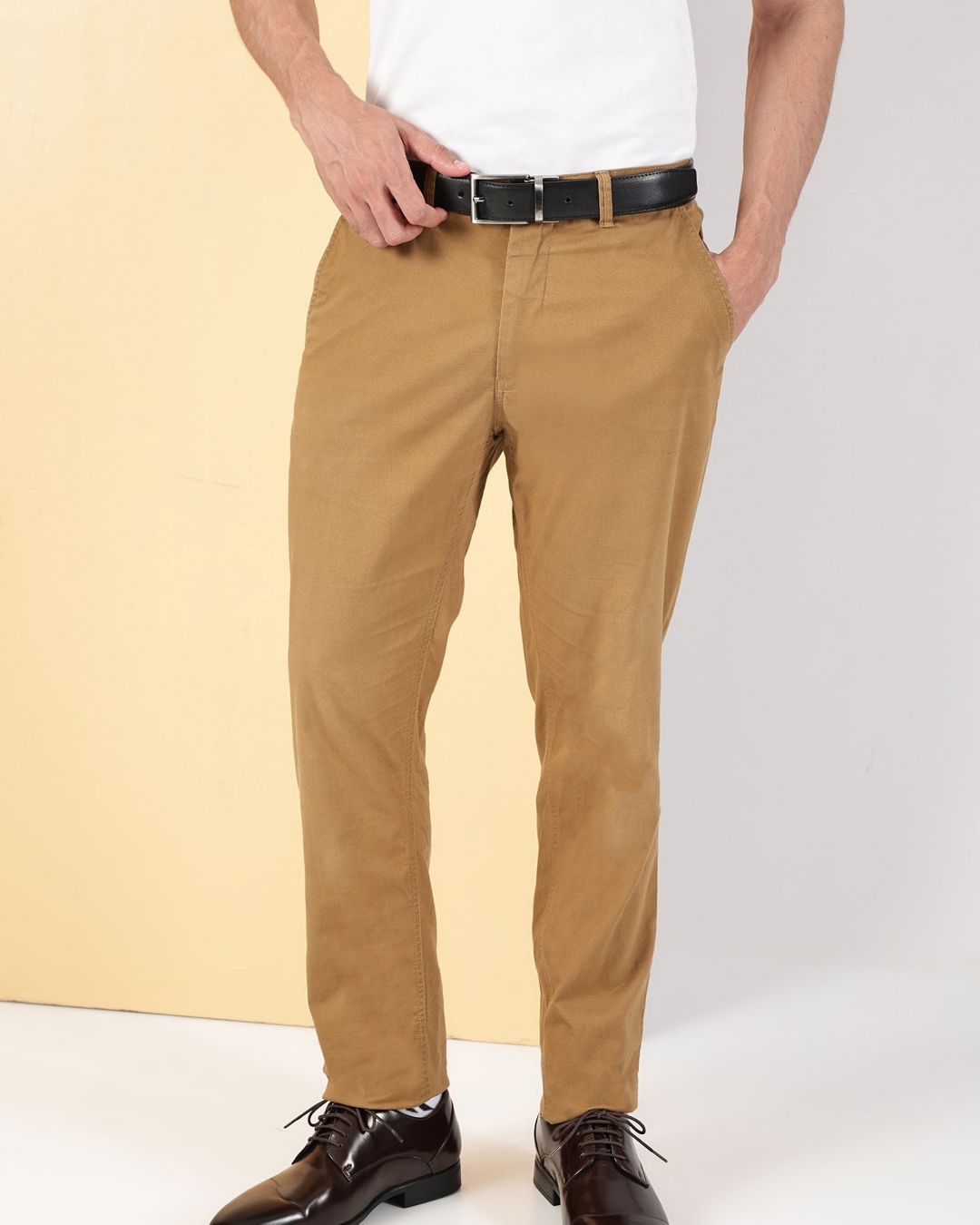A person wearing slim-fit tan chinos with a black belt, paired with a white t-shirt and dark brown dress shoes, shown from the waist down against a split beige and white studio background.