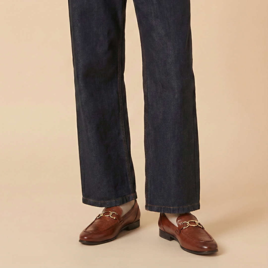A person wearing dark blue denim jeans and brown leather loafers with gold-tone horsebit details, standing against a solid beige background.
