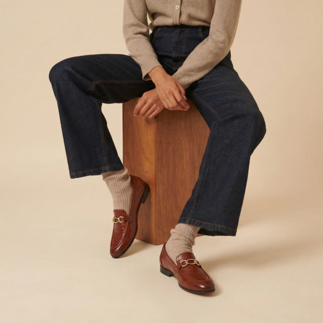 Person wearing brown loafers, dark jeans, and a light sweater sitting on a wooden block against a beige background.